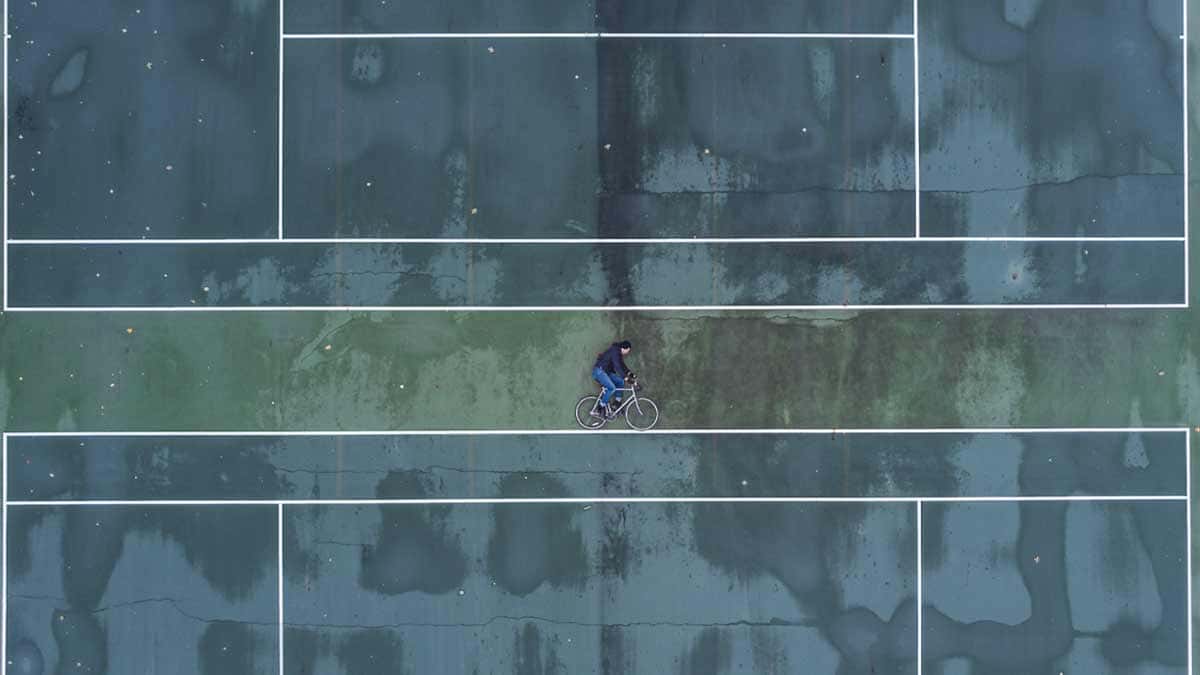 アイキャッチ画像｜上空撮影によるテニスコートに横たわり、自転車に乗っているように見せているだまし絵｜デザイン発注の教科書｜